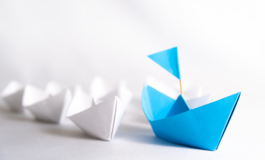 A blue paper boat with flag and white paper boats without flag, the former being the leader of the pack