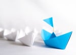 A blue paper boat with flag and white paper boats without flag, the former being the leader of the pack